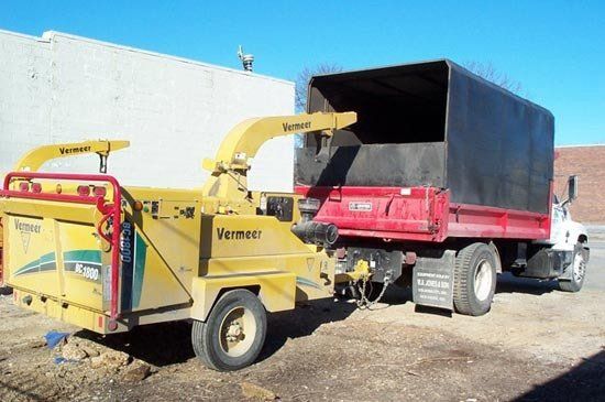 A yellow Vermeer wood chipper towed by a red dump truck parked against a white wall under a clear blue sky.