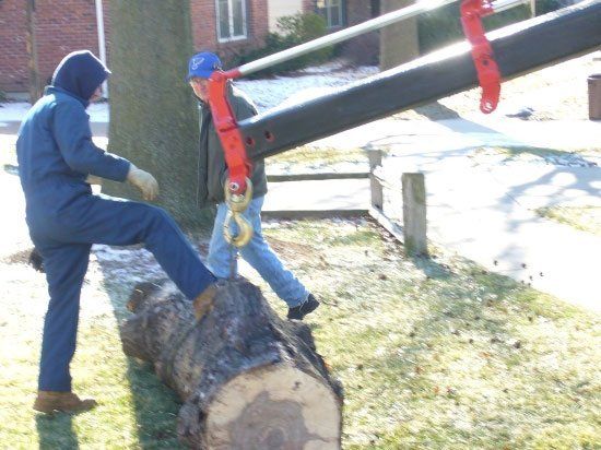 Two people maneuvering a heavy log with a crane attachment on a sunny, grassy day.