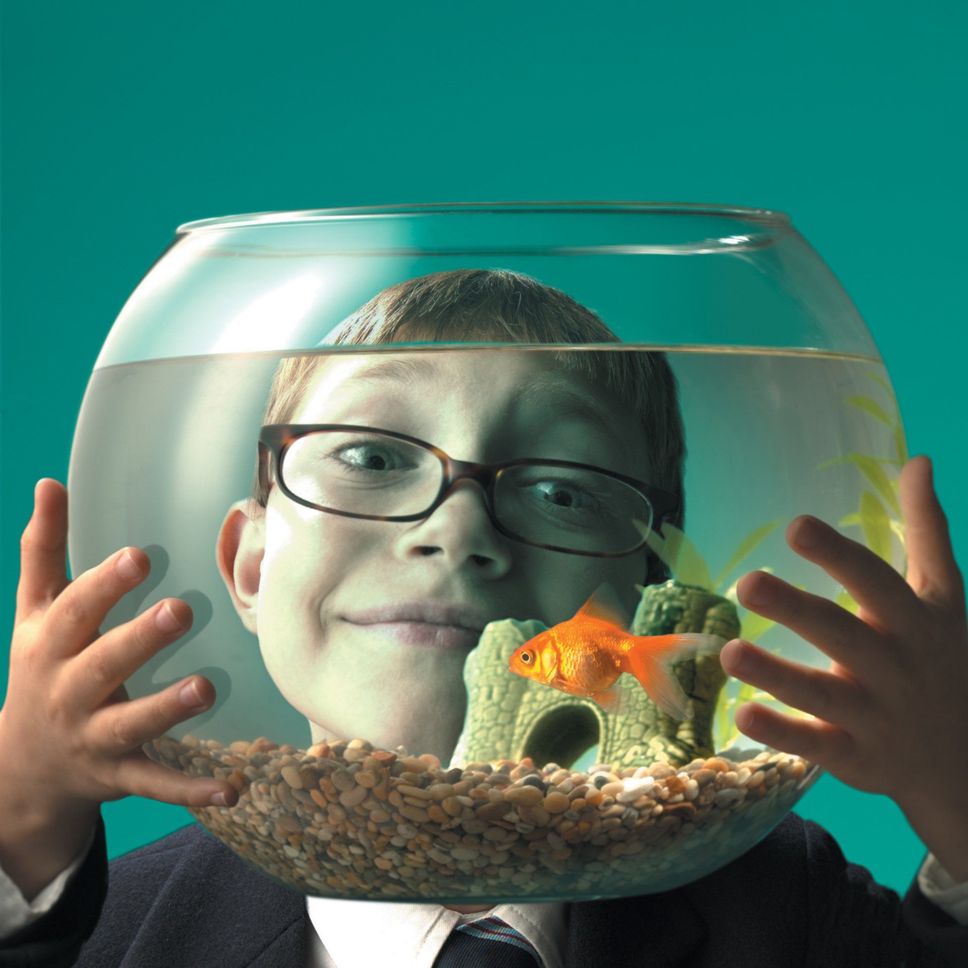A boy with glasses is holding a fish bowl in front of his face