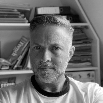 A man with a beard is taking a selfie in front of a bookshelf.