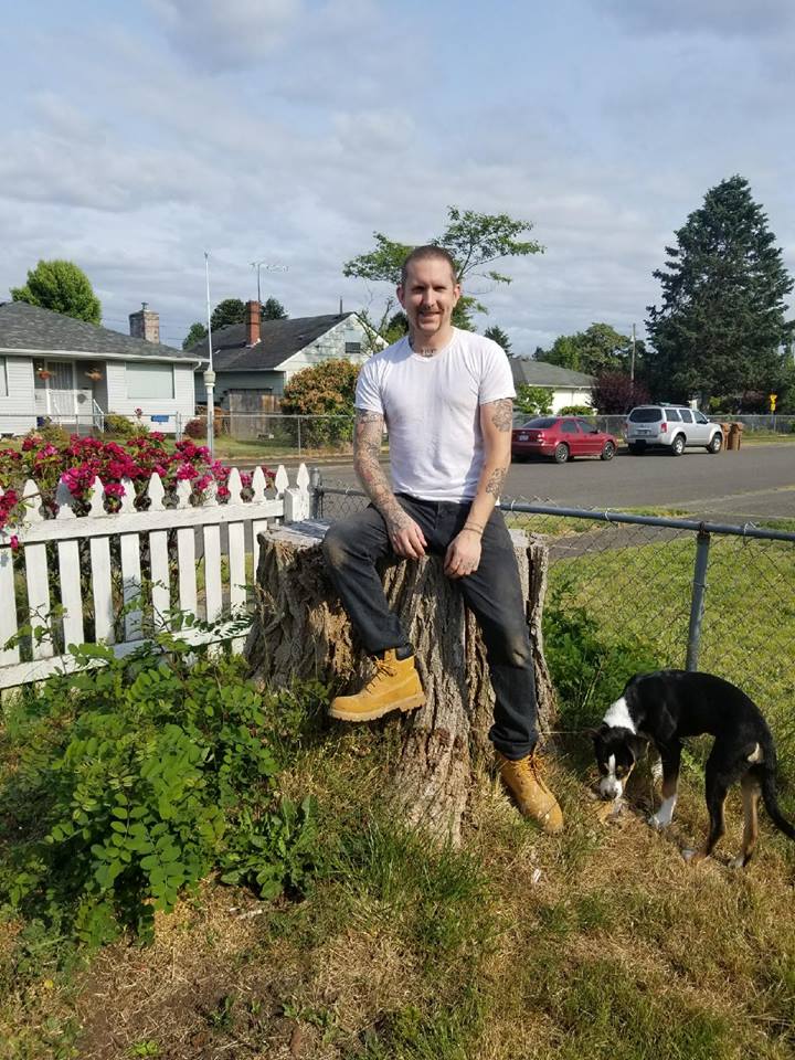 Owner Chris Wright sitting casually in the back yard on a tree stump sitting next to his black and white dog.