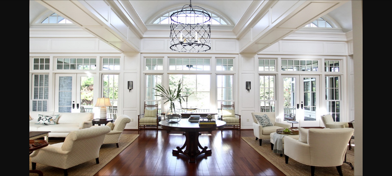 A large lounge with cream sofas and chairs, split by a dining table on a wooden floor, leading to long glass windows with patio doors