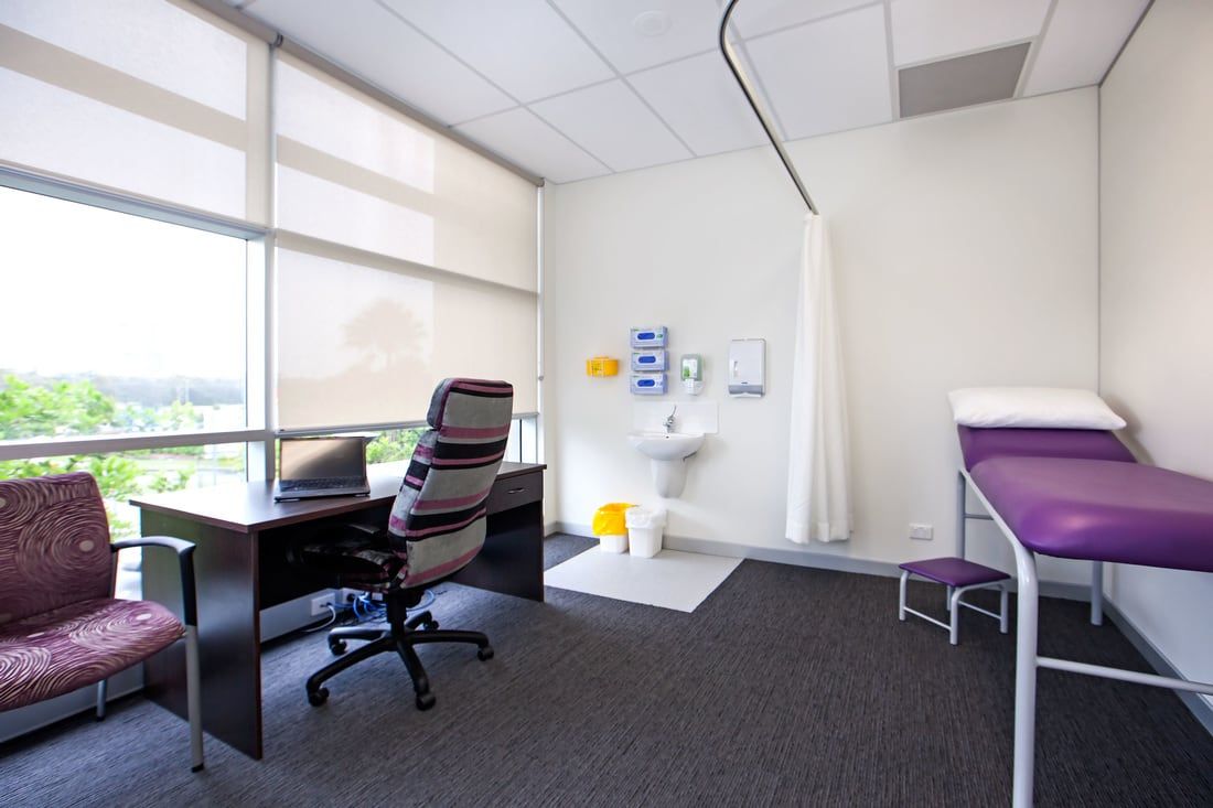 Exam room with exam table, desk, chair, and medical equipment by a window.