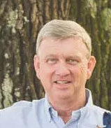 A man in a blue shirt is smiling for the camera in front of trees.