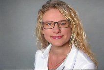 Frau mit blonden Haaren und Brille lächelt, trägt ein weißes Hemd, grauer Hintergrund.