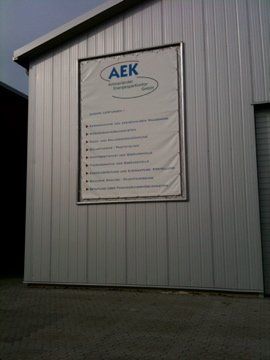 Schild an einem Metallgebäude mit den Dienstleistungen von „AEK“. Graue Außenverkleidung.