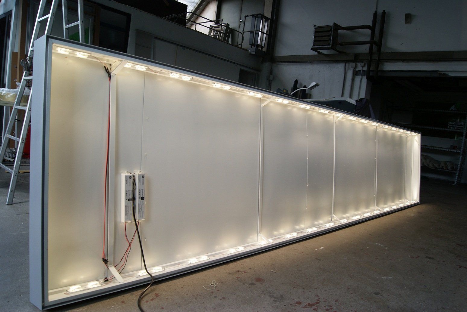 Großes, rechteckiges Leuchtschild mit LED-Leuchten an den Rändern. Weiße Paneele und Metallrahmen in einer Werkstattumgebung.