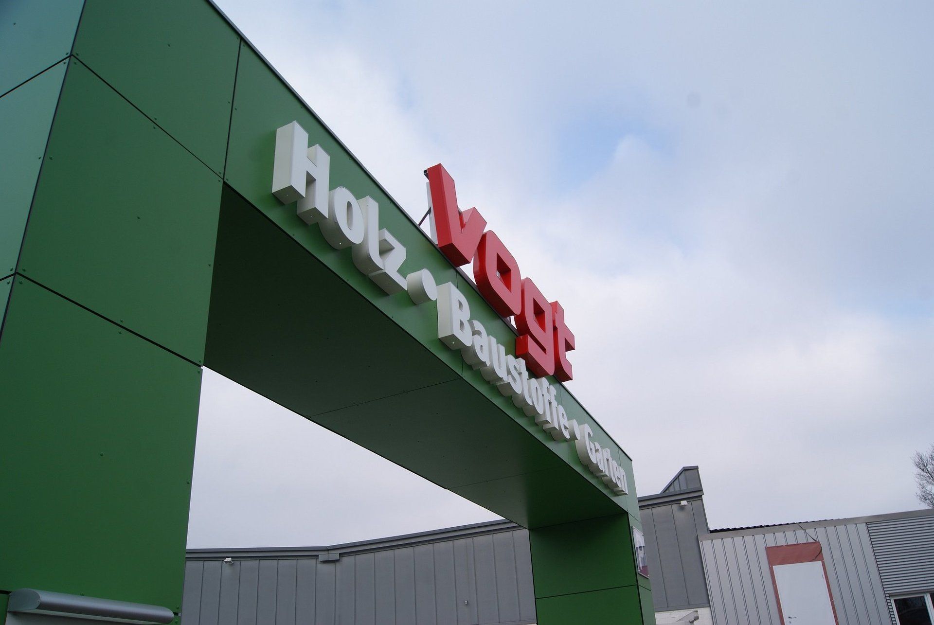 Grüner Eingangstorbogen mit rot-weißem Schild: „Holz Vogt Bauzentrum“. Bewölkter Himmel.