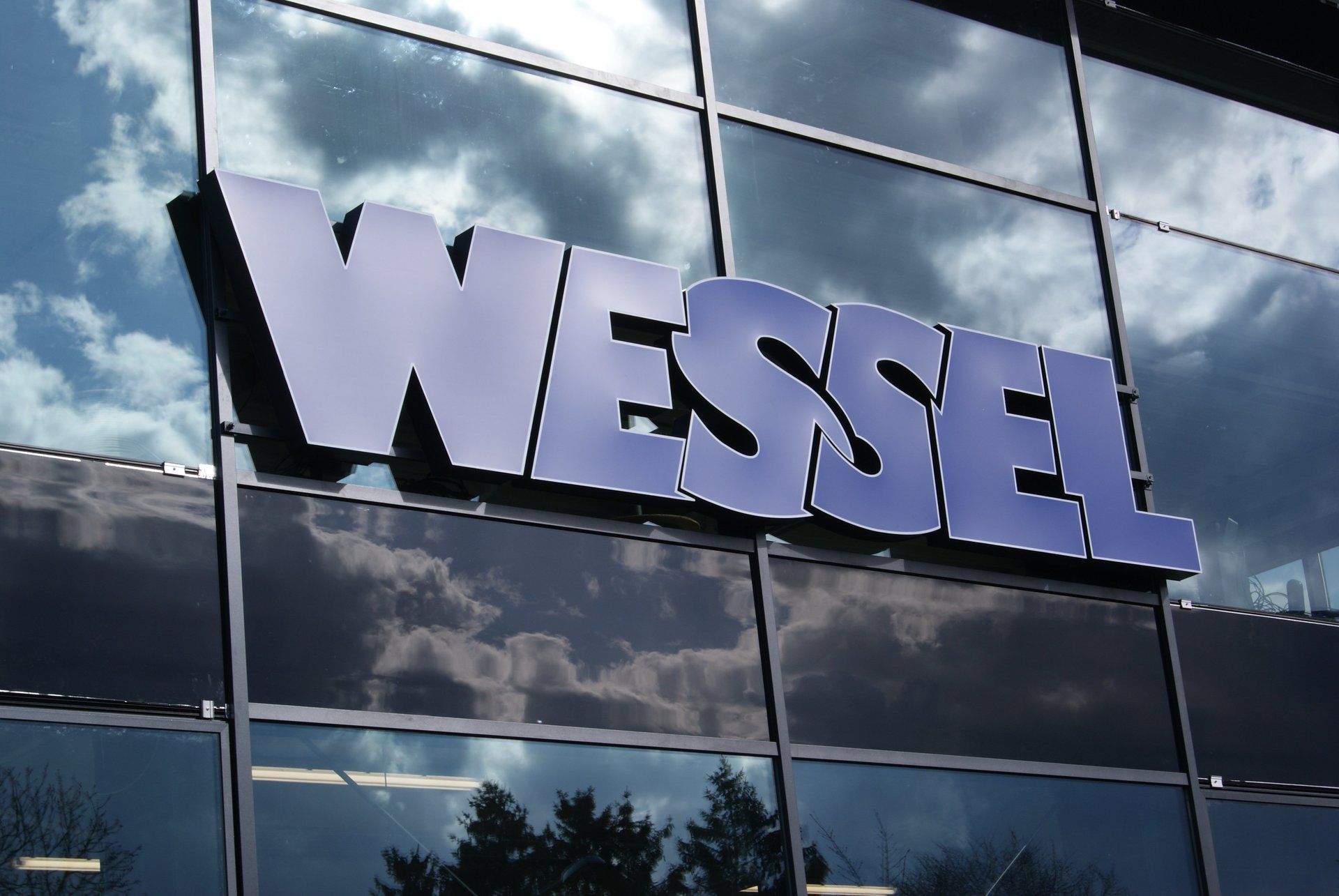 „WESSEL“-Schriftzug in Blau auf Glasfassade; Himmelsspiegelung sichtbar.