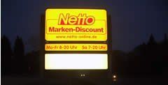 Beleuchtetes Netto-Supermarktschild bei Nacht; gelbes und rotes Logo, Öffnungszeiten angegeben.
