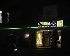 Außenansicht des medizinisch-ästhetischen Instituts „GESUNDSCHÖN“ bei Nacht, beleuchtetes Schild mit grünen Akzenten.