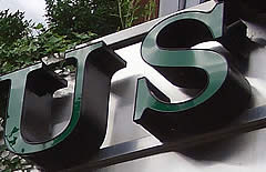 Schild mit den Buchstaben „UFS“ in Dunkelgrün, montiert auf einer silbernen Oberfläche.