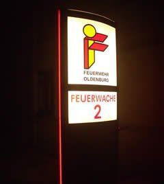 Feuerwehrschild, Logo „Feuerwehr Oldenburg“, Schriftzug „Feuerwache 2“, rot-weiß beleuchtet auf schwarzem Pfeiler.