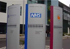 Drei große, silberne Schilder mit Firmenlogos und Text, eines mit NHS-Logo, im Freien.