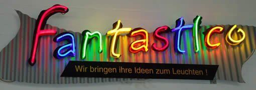 Leuchtreklame: „Fantastico“ in Regenbogenfarben mit deutschem Slogan auf gestreiftem Hintergrund.