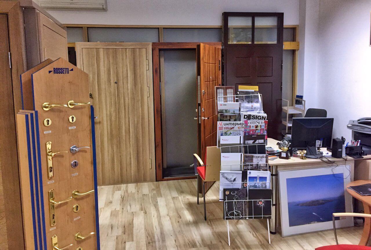 Showroom with various doors on display, a door handle display, and an office desk with promotional materials.