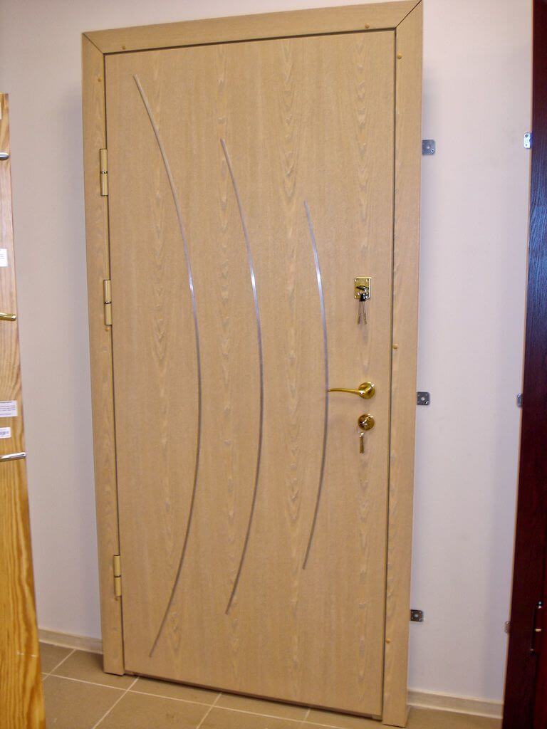 Tan wooden door with three curved silver inlays, gold hardware, and metal reinforcement plates on a white wall.