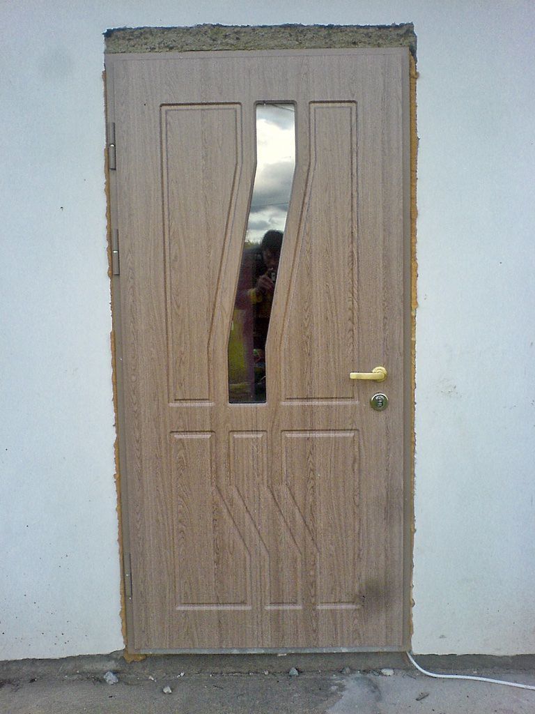Tan door with angled glass panel, set in a white wall with unfinished edges.