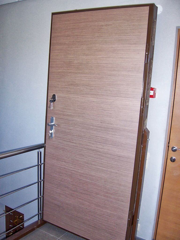 A closed light brown wooden door with a silver handle and multiple locks, beside a metal railing.