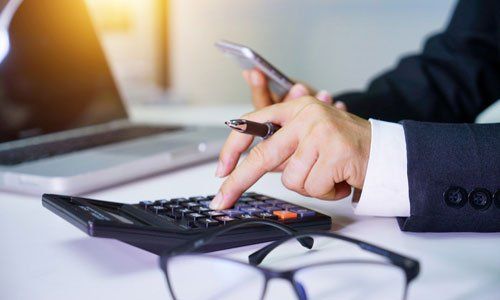 Hands using a calculator beside a laptop and phone in an office setting