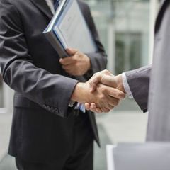 Two businesspeople shaking hands; one holds a blue folder in an office setting