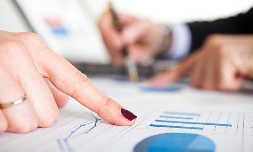 Hand pointing at a financial chart on paper during a meeting, with blurred coworkers in the background