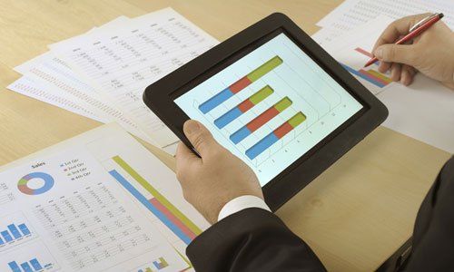 Person using a tablet to view colorful bar charts while writing on papers at a desk