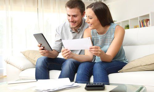 Two people on a couch reviewing bills with a tablet and paper invoices in a bright living room