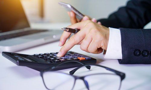 Hands using a calculator with a pen and smartphone on a desk near eyeglasses.