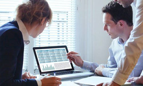 Three coworkers reviewing charts on a laptop in a bright office meeting room