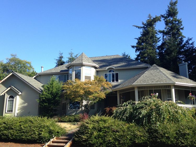 Large gray suburban house with a porch, surrounded by green trees and shrubs under a blue sky
