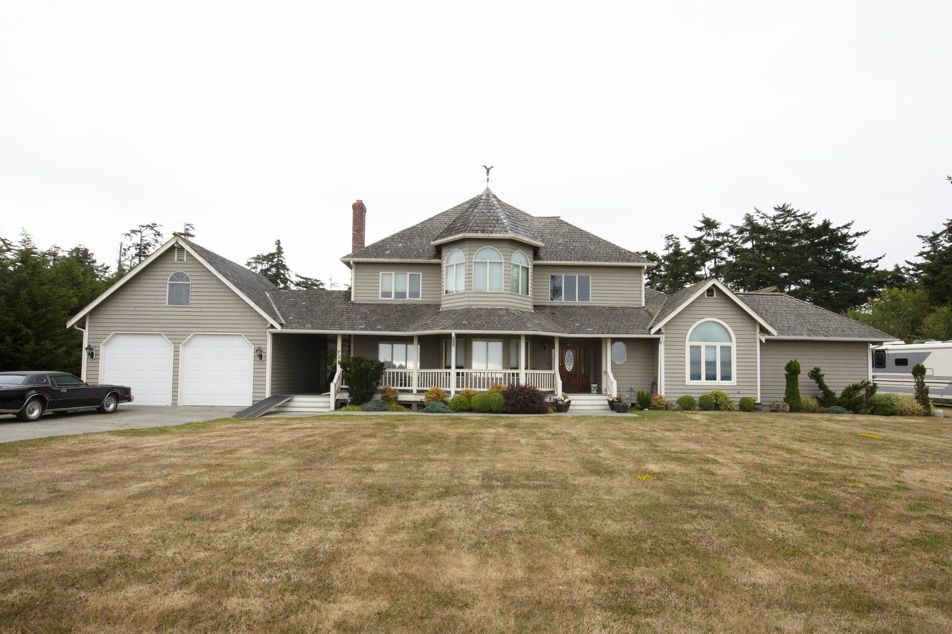 Large gray suburban house with a front porch, double garage, and dry front lawn under an overcast sky