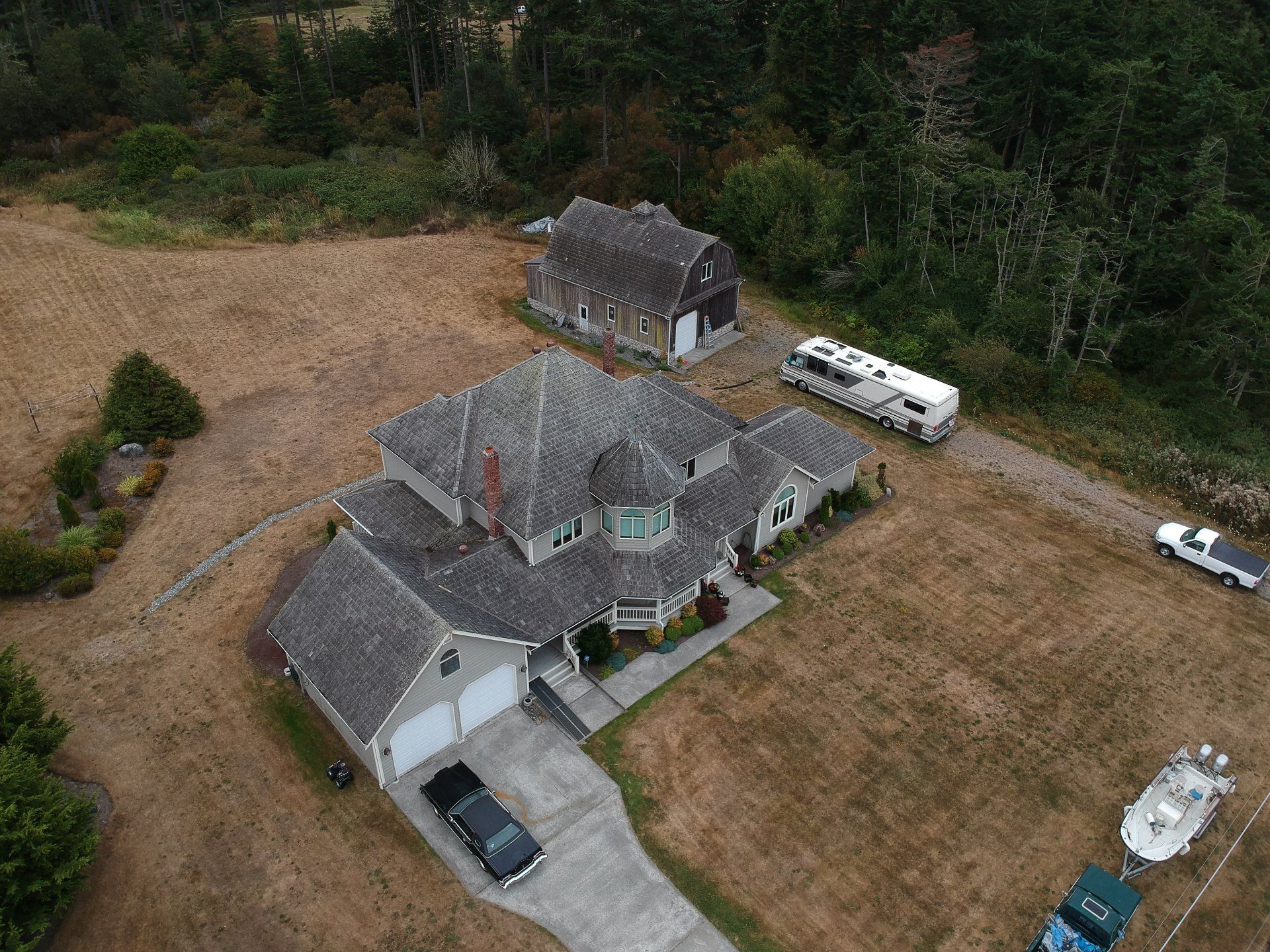 Aerial view of a gray house with a driveway, detached shed, parked vehicles, and a boat beside a wooded yard.