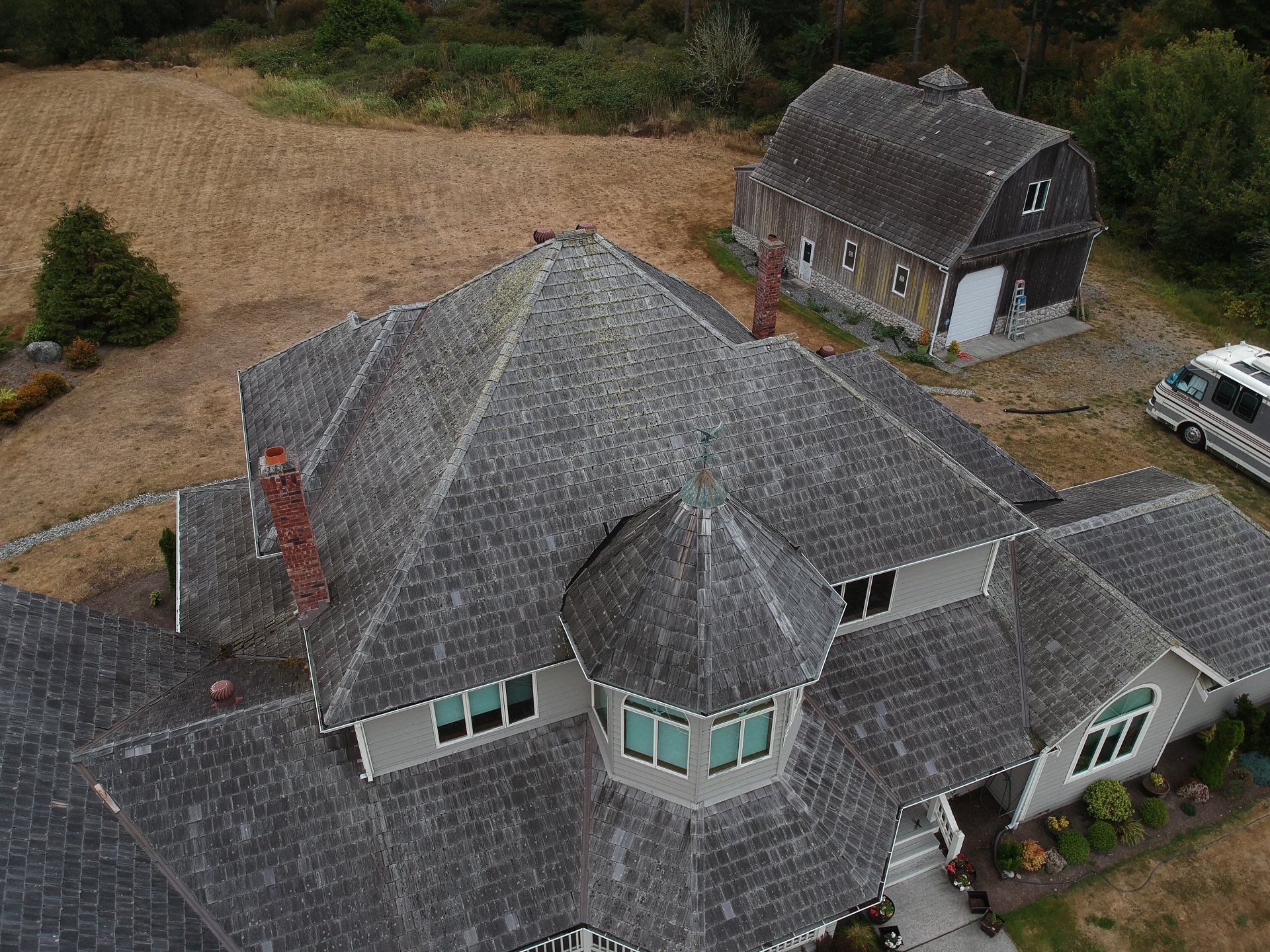 Aerial view of a gray-roofed house with dormers and a detached garage in a gravel yard