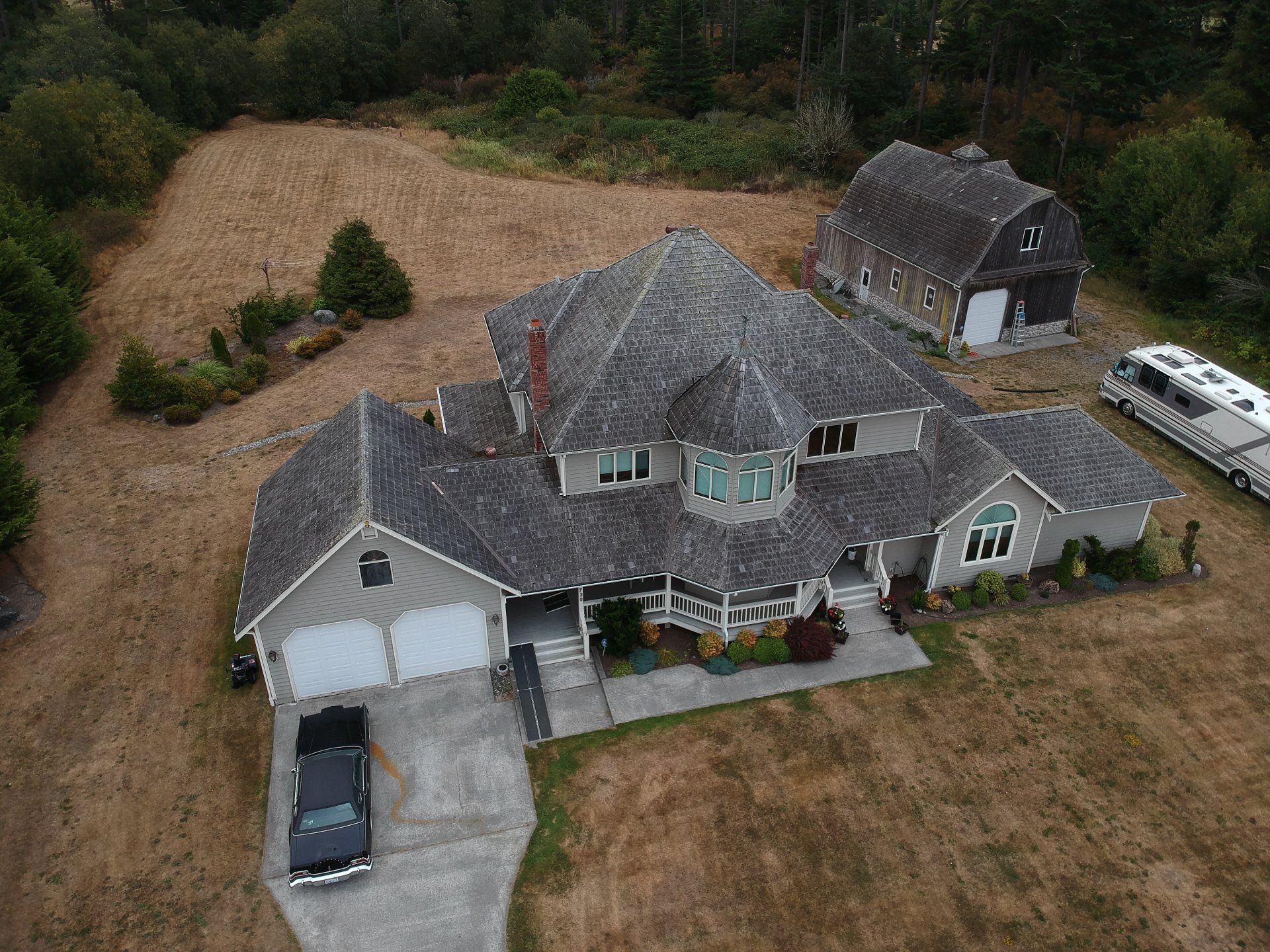Aerial view of a large gray house with a driveway, garage, and surrounding trees on a dry lawn