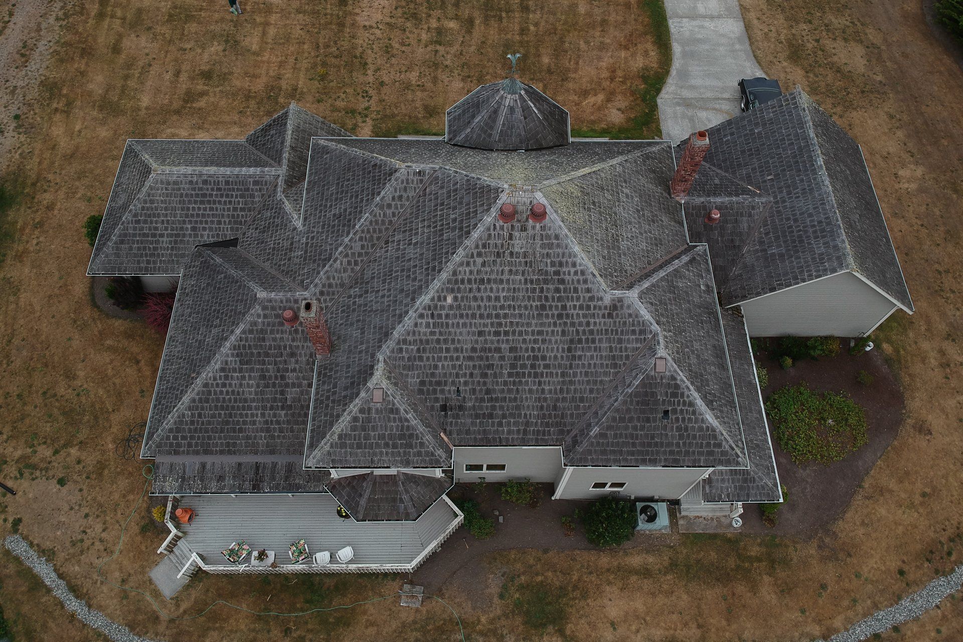 Aerial view of a gray-roofed house with multiple gables and a brown lawn surrounding it
