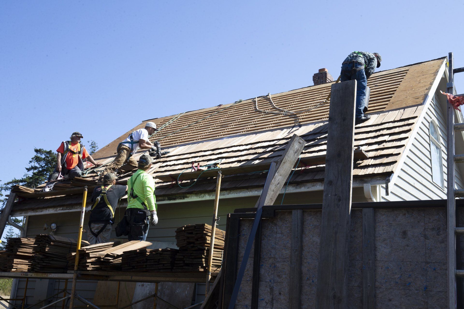 Roofers working on a house roof beside a tall chimney and a white-sided wall