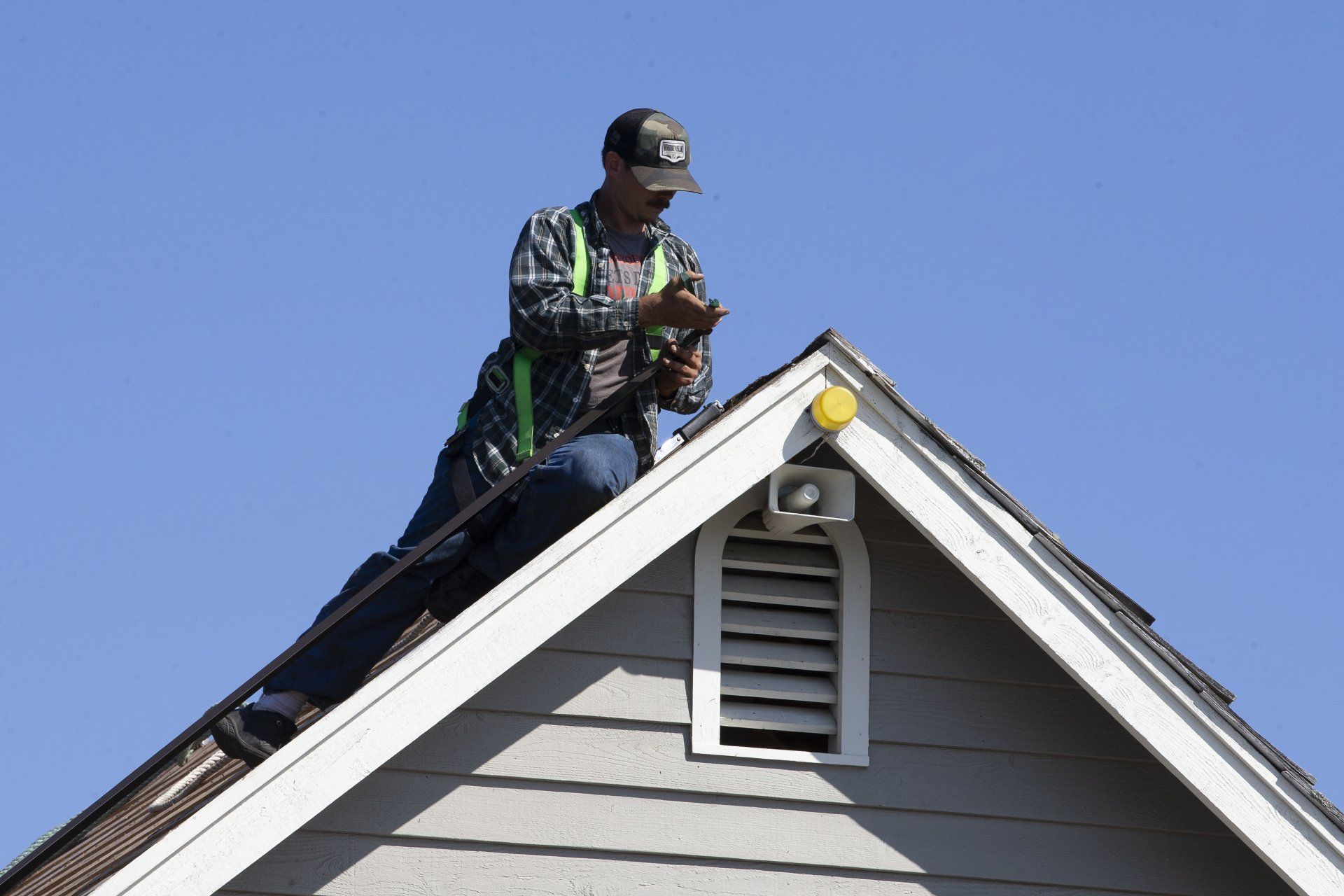 Worker on a rooftop repairing shingles against a clear blue sky.