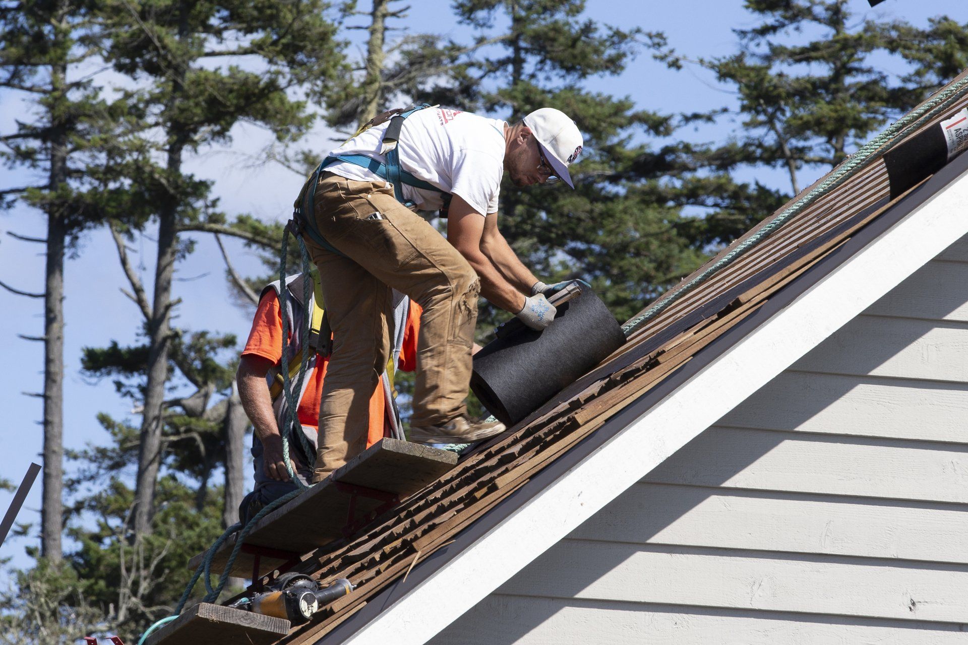 Workers repairing a shingled roof on a house, with one carrying a black roofing roll.