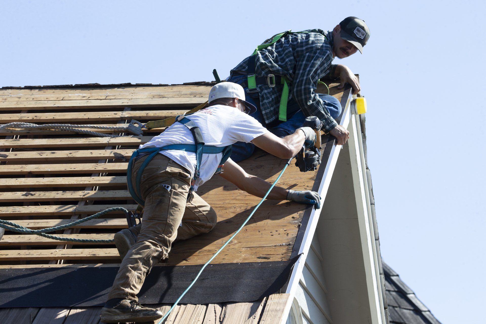 Two workers on a roof securing metal roofing panels with tools and safety gear