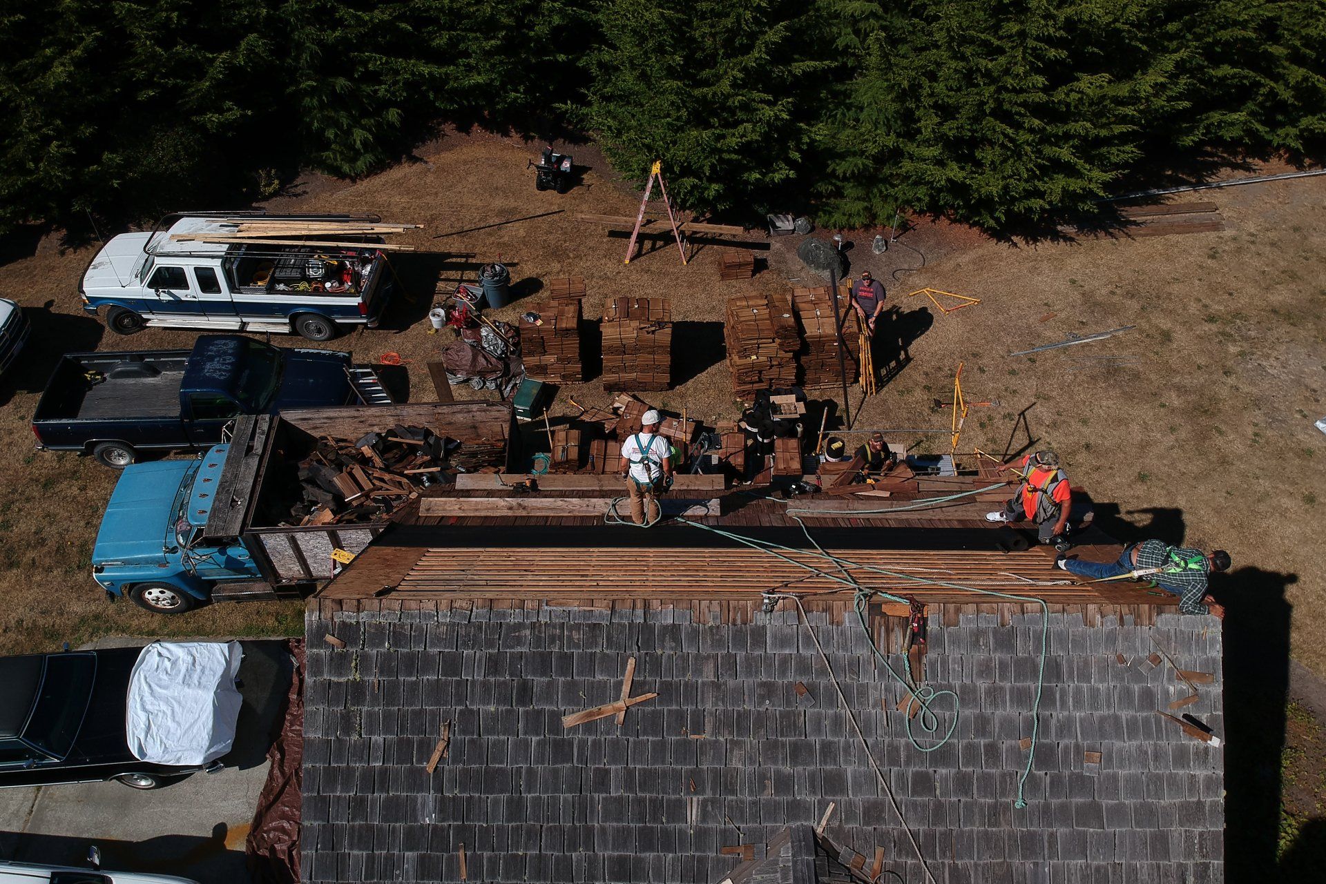 Aerial view of workers on a roof beside a white truck and stacked lumber in a wooded area