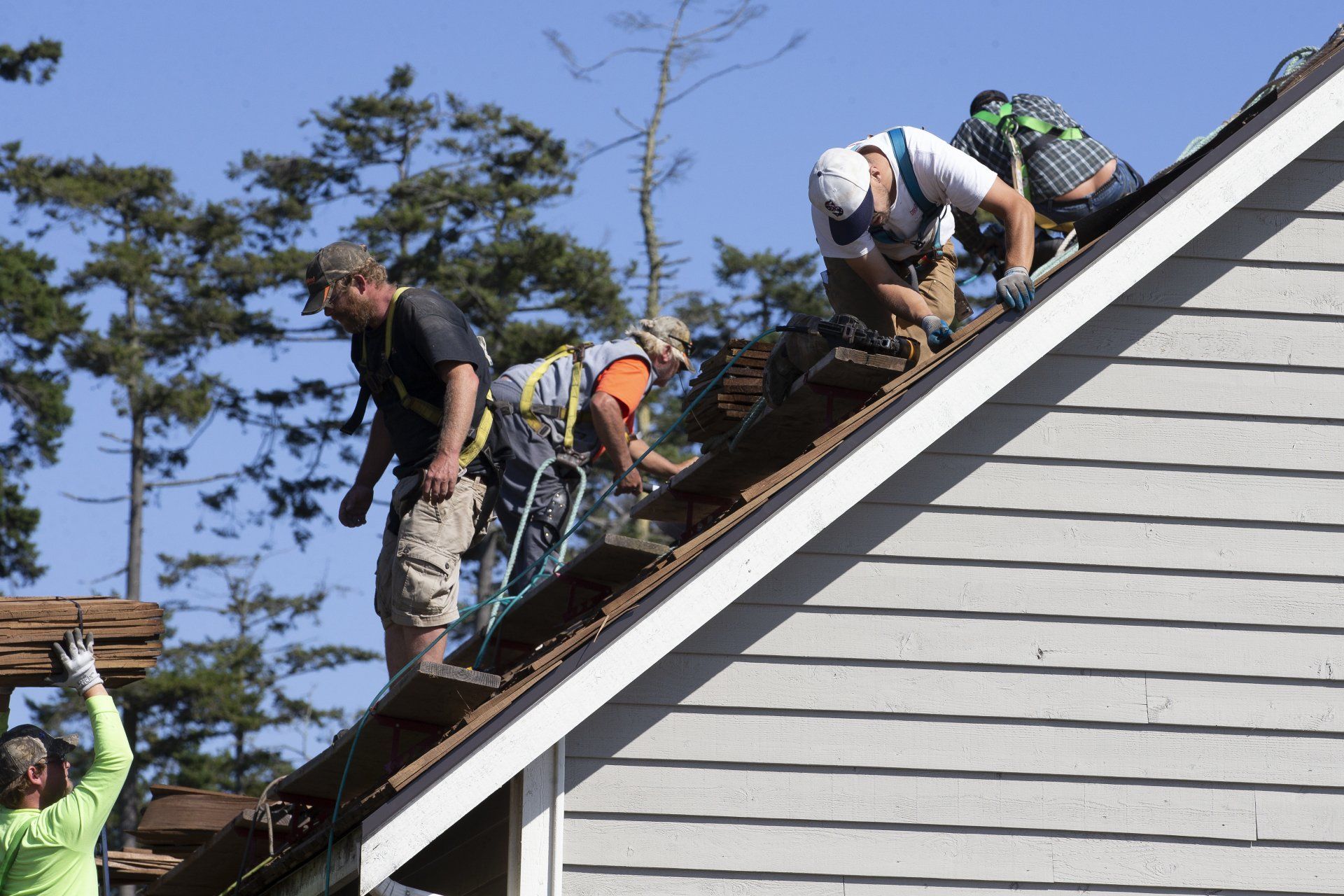 Workers repairing a gray shingled roof, several crouched along the ridge line.