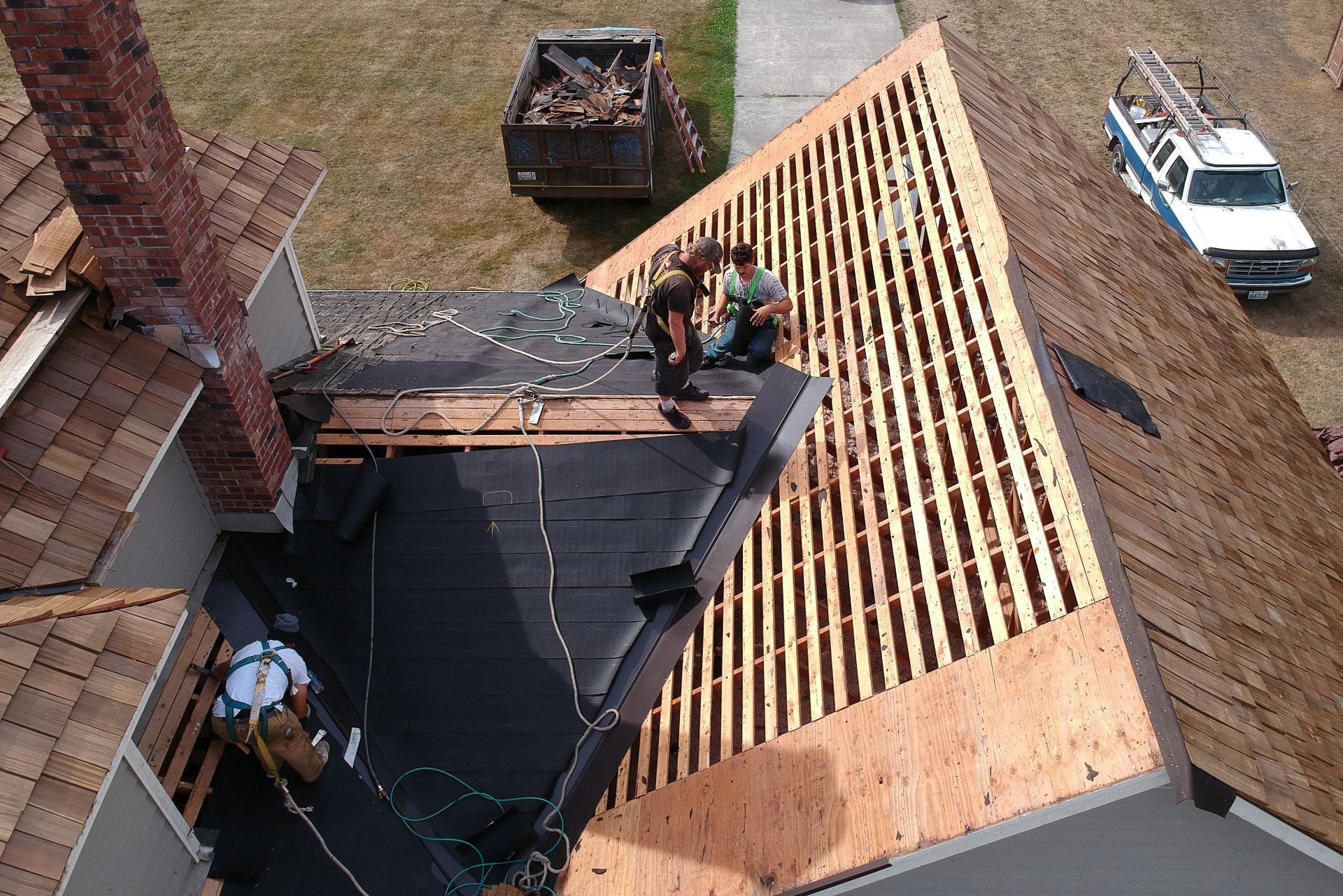 Workers installing a shingle roof on a house, with lumber, ladders, and a pickup truck nearby.