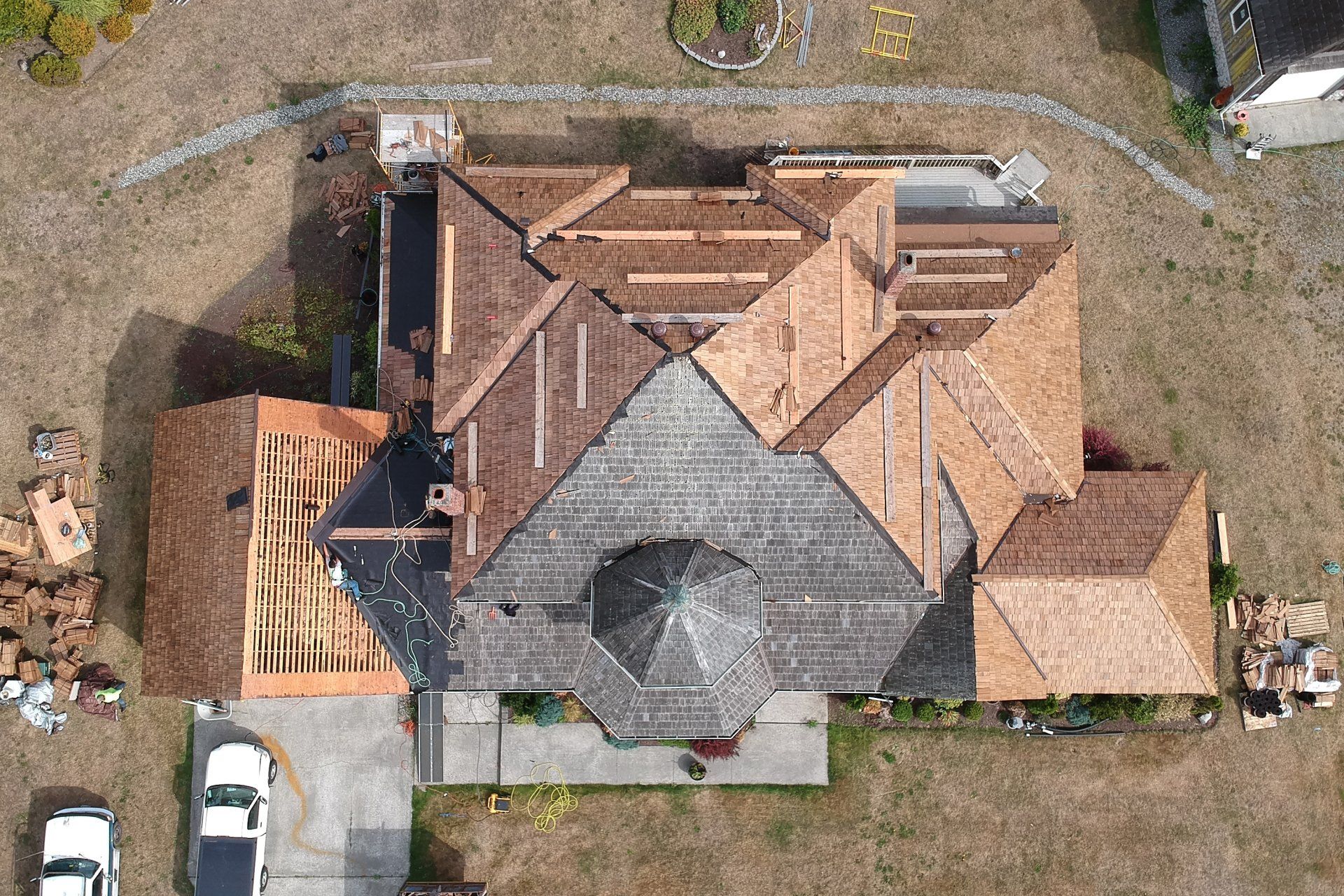 Aerial view of a house with a gray central roof and tan roof sections, surrounded by yard and driveway.