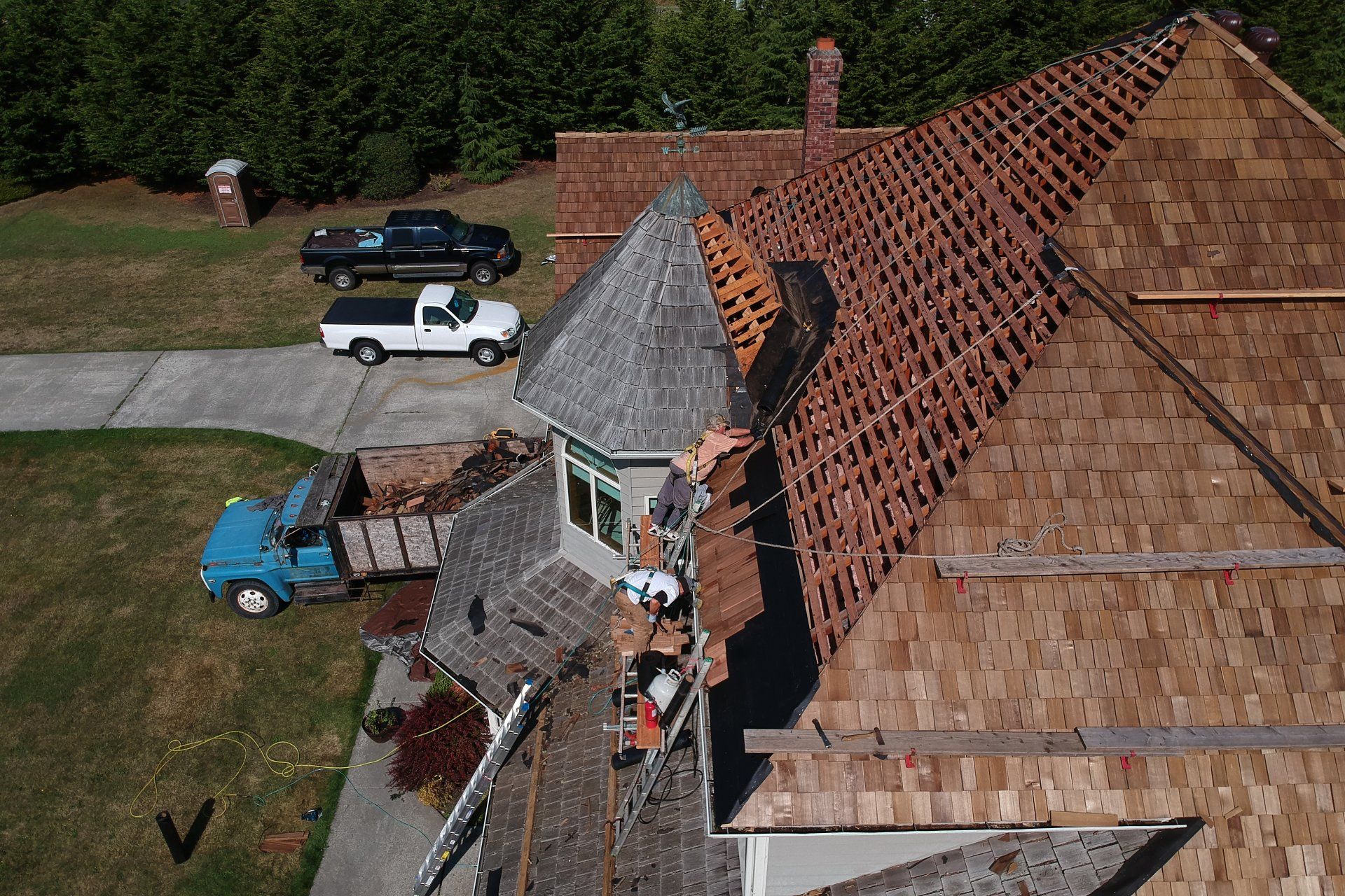 Damaged roof on a house with workers repairing it, surrounded by a driveway and parked vehicles.