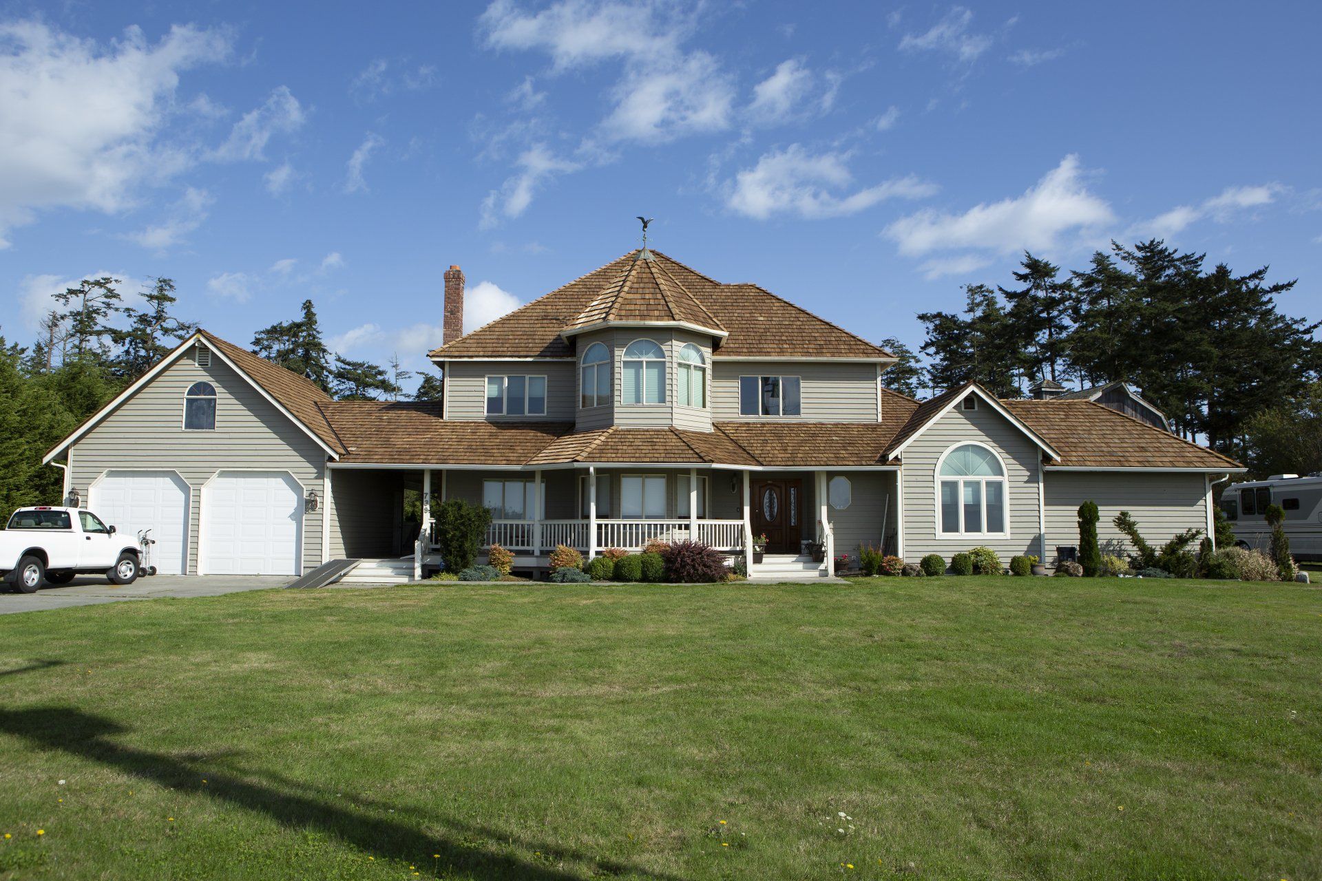 Large suburban house with a wraparound porch, white garages, and a wide green lawn under a blue sky