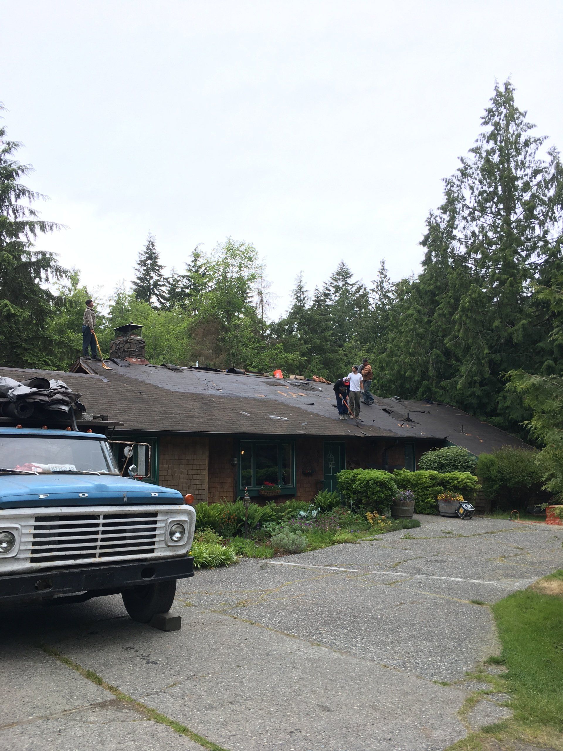 Blue vintage truck parked by a house with people on the roof and a gravel driveway.