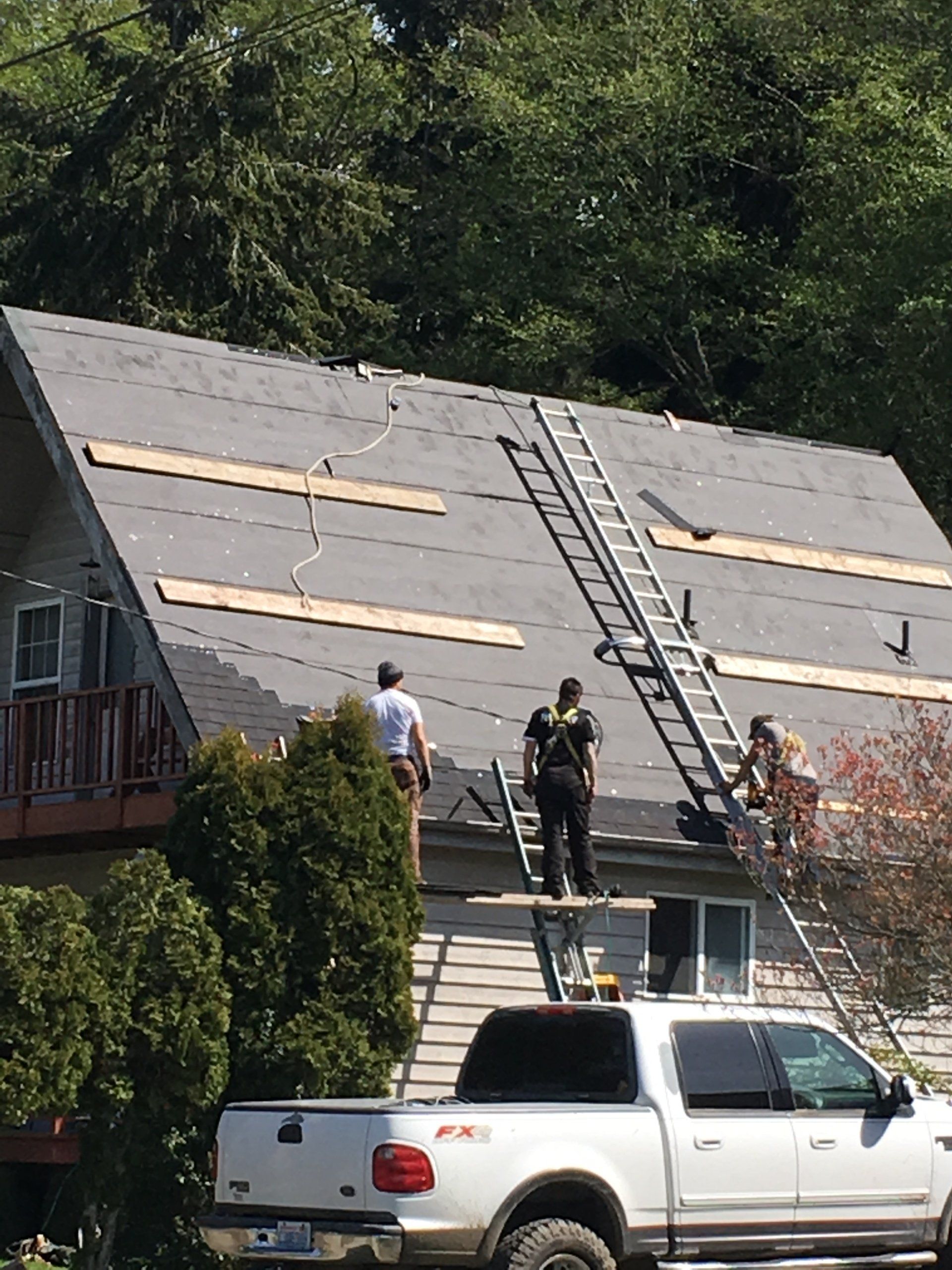 Workers on a steep roof installing shingles with two ladders; truck parked below.