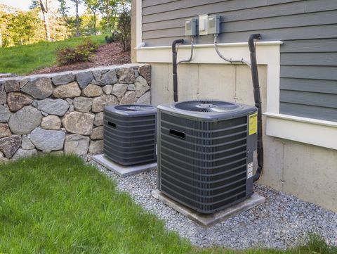 Two air conditioners are sitting outside of a house next to a stone wall.
