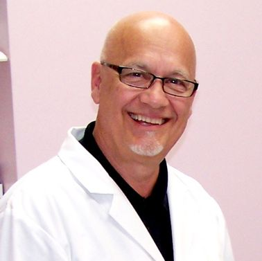 A bald man wearing glasses and a white lab coat smiles for the camera